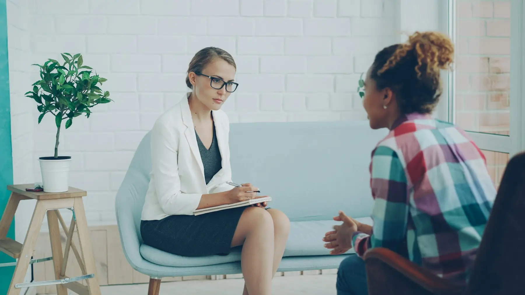 Psicóloga conversando com uma paciente em um consultório claro, com plantas ao fundo